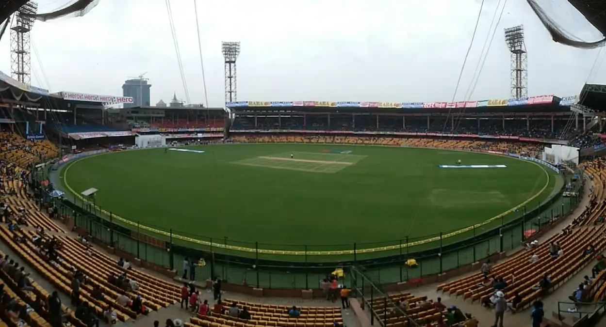 A wide shot of the M. Chinnaswamy Stadium in Bangalore, India, before the start of a day cricket match. The stadium is partially filled with spectators entering the stands, and the green field and pitch are visible in the center.